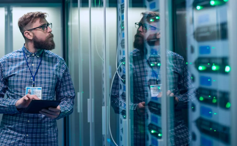 A man holding a tablet while standing in a server room, surrounded by rows of servers and networking equipment.